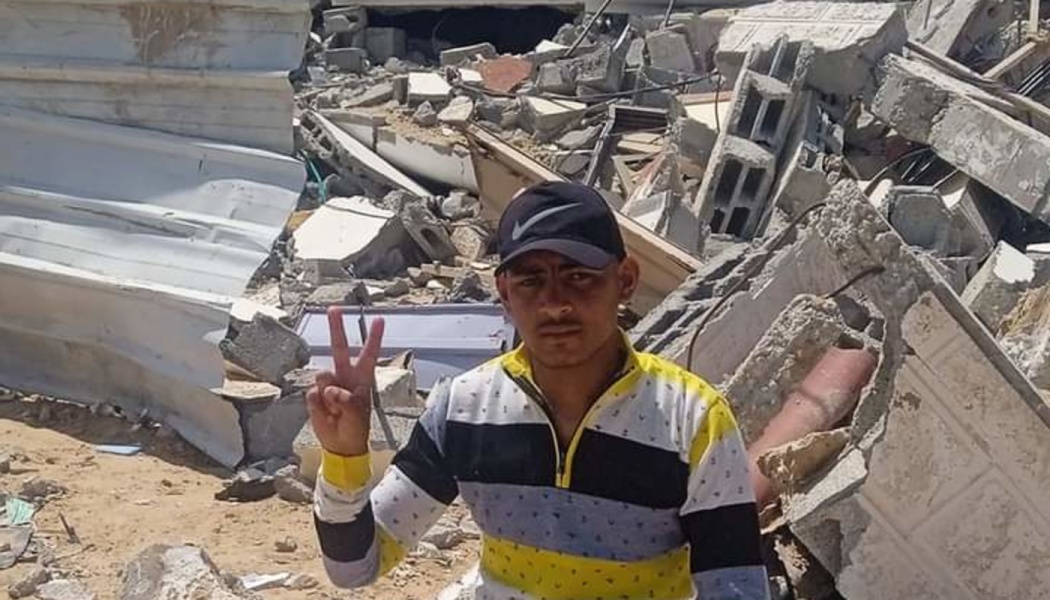 A young man in a baseball cap holds up a peace sign while standing amidst the rubble of a destroyed building.