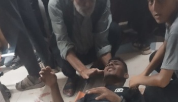 An injured young man lies on the floor in pain while an older man comforts him, surrounded by bystanders.
