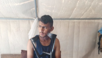 A young man in a blue tank top sitting inside a tent structure, holding a pair of black crutches.