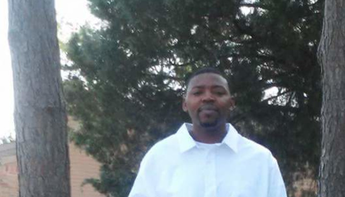 A Black man in a white collared shirt standing outdoors between pine trees.