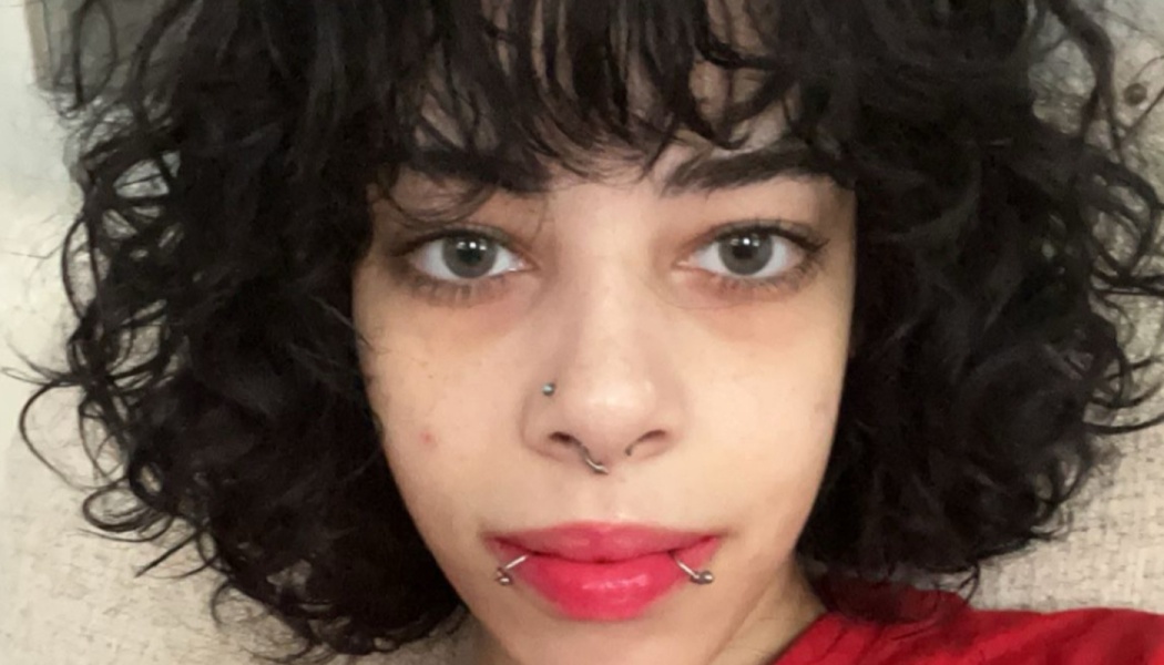 Close-up portrait of a person with dark curly hair, light eyes, facial piercings, and red lipstick.
