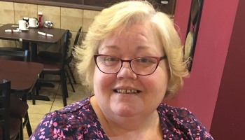 A smiling woman with short blonde hair and red glasses wearing a patterned top in a restaurant.