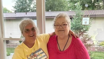 Two smiling senior women with glasses embrace indoors, one wearing yellow and the other wearing pink.