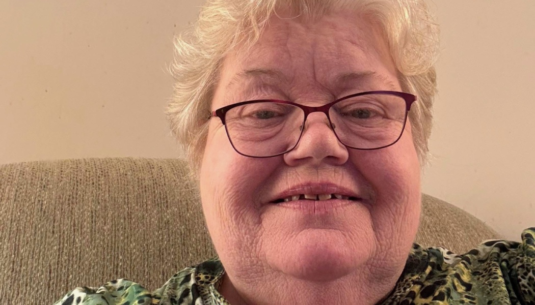 An elderly woman with short blonde hair and red glasses smiling warmly at the camera.