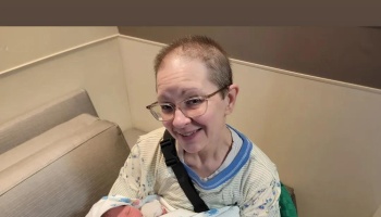 A smiling woman with short hair and glasses tenderly holding a swaddled newborn baby.