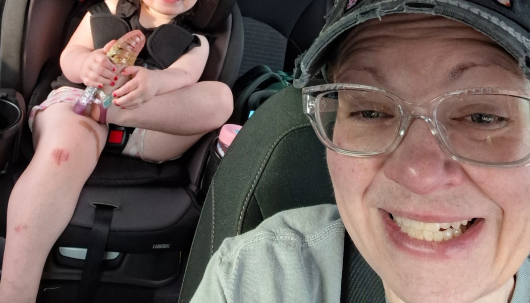 A smiling adult takes a car selfie, with a young child in a car seat holding a glittery toy behind them.