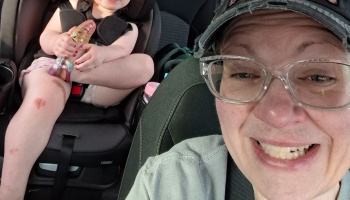 A smiling adult takes a car selfie, with a young child in a car seat holding a glittery toy behind them.