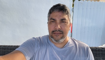 Middle-aged man with a salt-and-pepper beard wearing a gray t-shirt, sitting outdoors in front of a white wall.