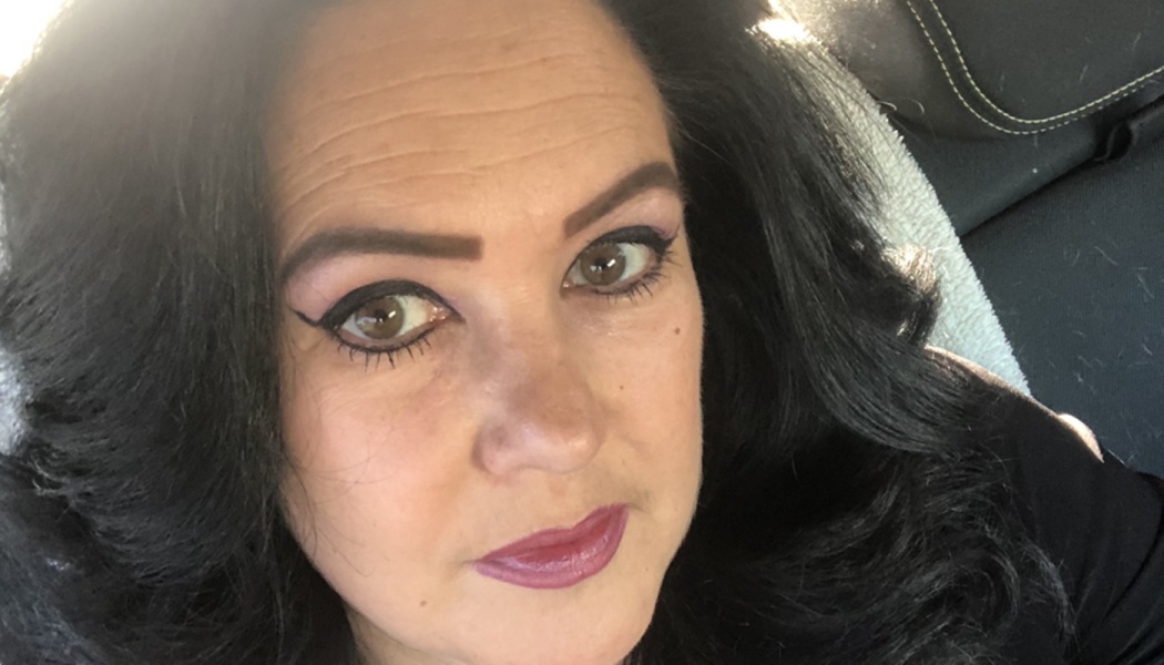 Close-up portrait of a woman with dark wavy hair, brown eyes, winged eyeliner, and berry lipstick.