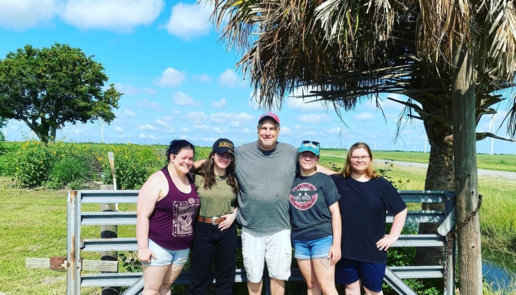 A smiling family of five poses together outdoors in front of a metal gate on a sunny day.