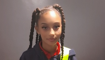 Young girl with long braided pigtails, a small heart under her eye, wearing a high-visibility vest.