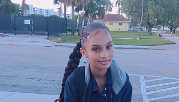 A smiling young girl with a long braided ponytail outdoors wearing a navy blue jacket.
