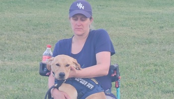 A woman sitting outdoors holds a yellow service dog wearing a vest that reads "SERVICE DOG DO NOT PET".