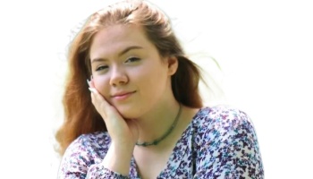 Young woman with long brown hair wearing a floral top, resting her hand on her cheek with a soft smile.