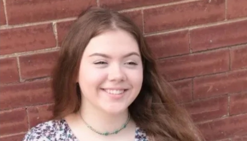 A smiling young woman with long brown hair and a green necklace against a red brick wall.