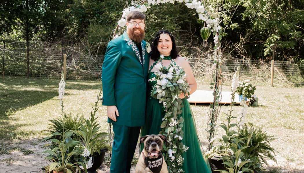 A smiling couple in matching emerald green wedding attire poses with their dog under a floral archway outdoors.