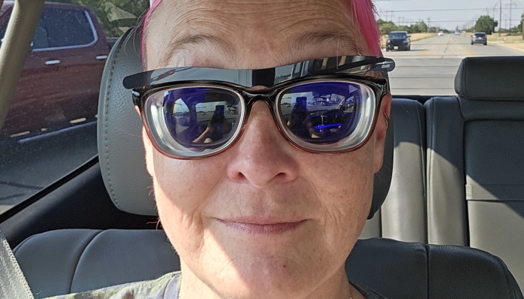 Close-up of a smiling person with bright pink hair wearing extremely thick-lensed glasses inside a car.