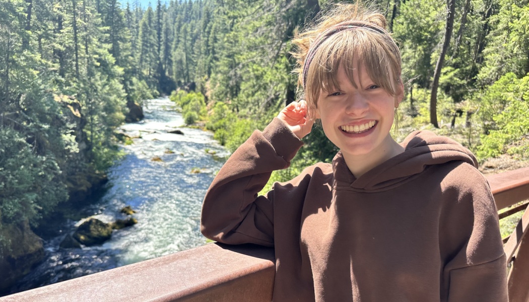 A smiling young woman in a brown hoodie stands on a bridge overlooking a scenic forest river.