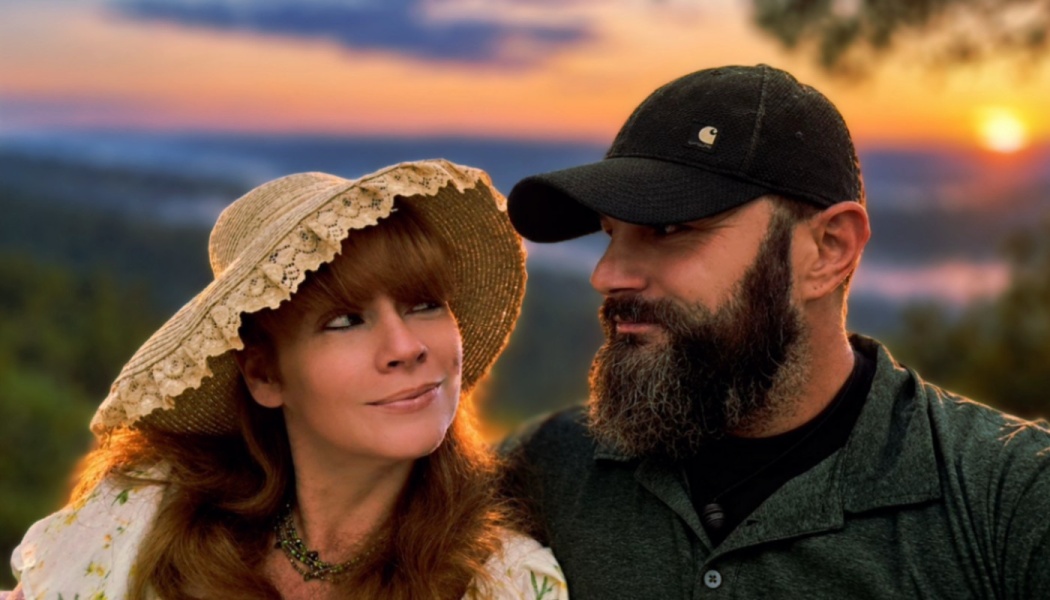 A woman in a straw hat and a bearded man in a cap gaze lovingly at each other with a bright sunset behind them.