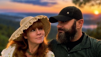 A woman in a straw hat and a bearded man in a cap gaze lovingly at each other with a bright sunset behind them.