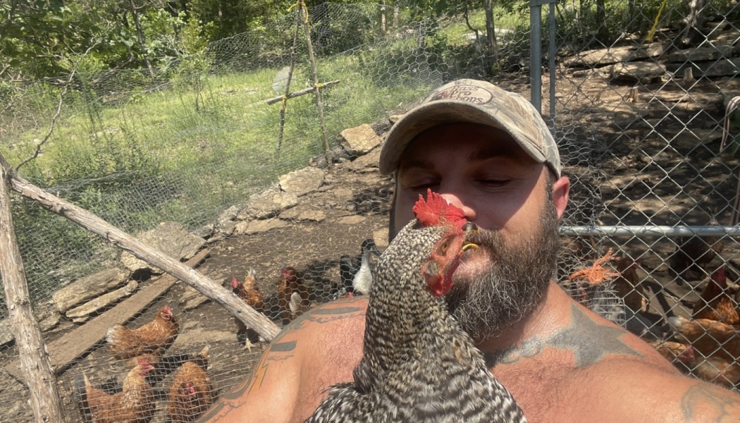 A bearded man in a camo hat poses close with a barred rock chicken inside an outdoor chicken coop.
