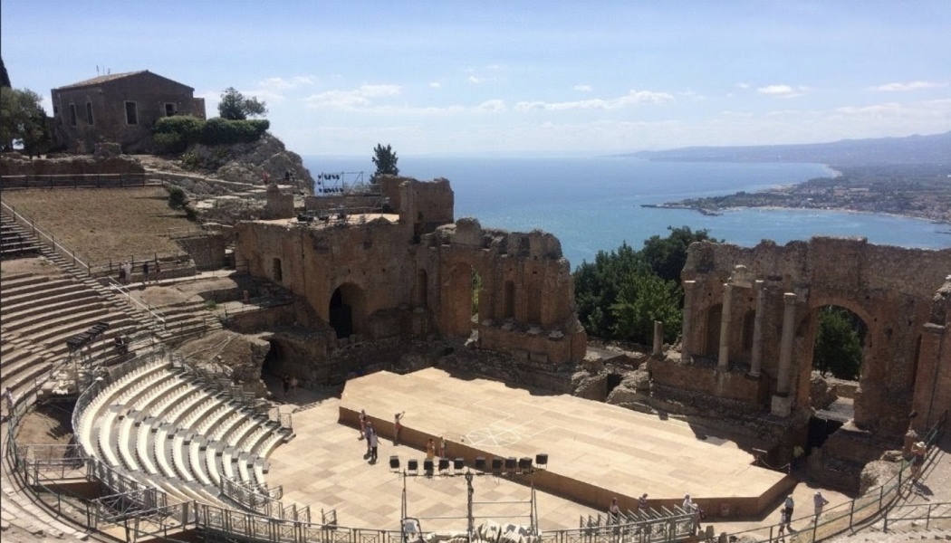 Ancient stone amphitheater with curved seating and a stage overlooking a scenic blue sea and coastline.
