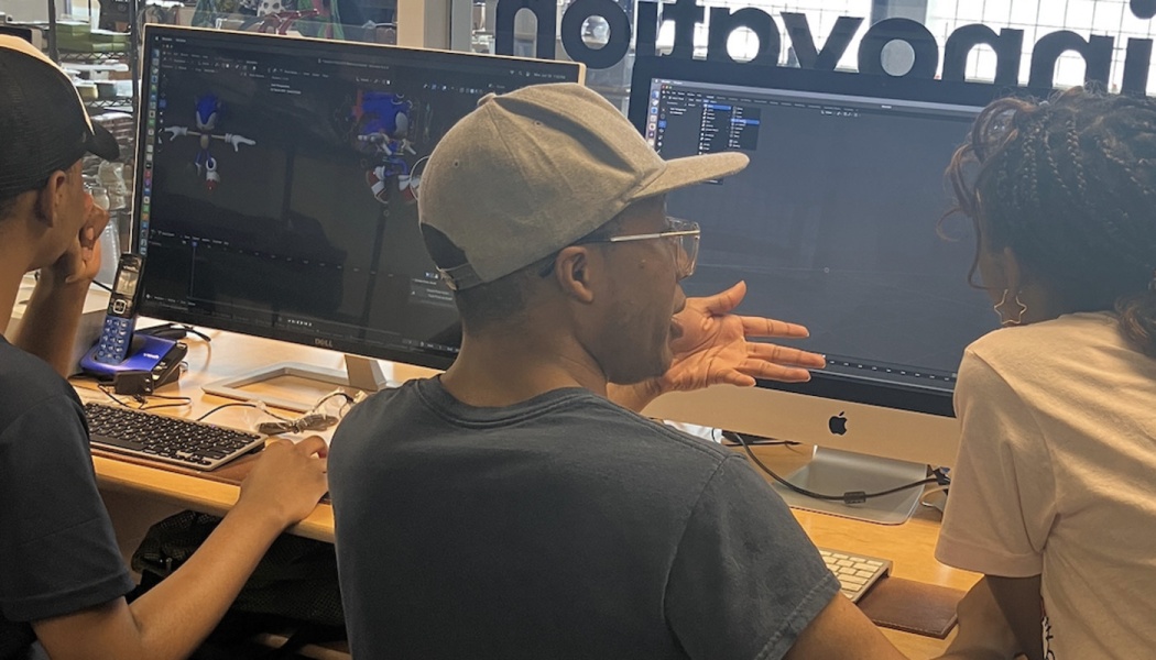 Three people collaborate around desktop computer monitors, discussing a 3D animation project.
