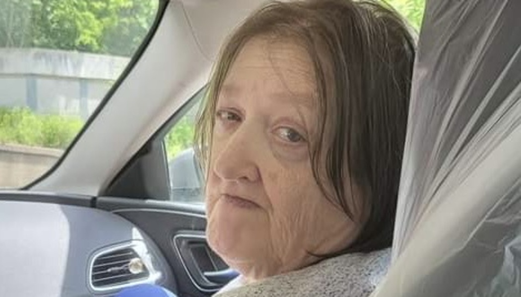 An elderly woman with brown hair sitting in a car, looking back toward the camera.