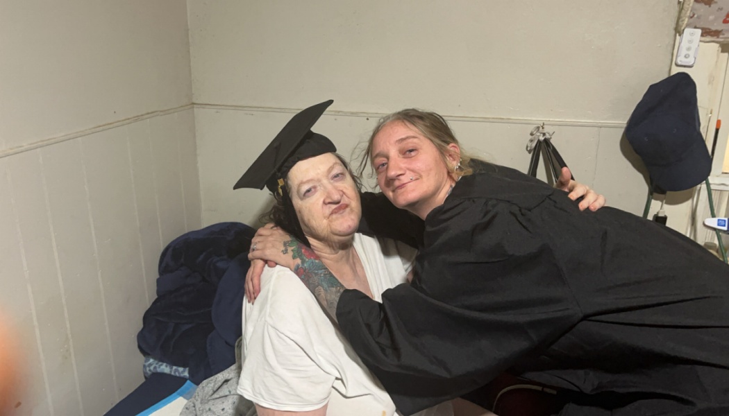 A woman in a black graduation gown warmly hugs an older woman wearing a graduation cap.