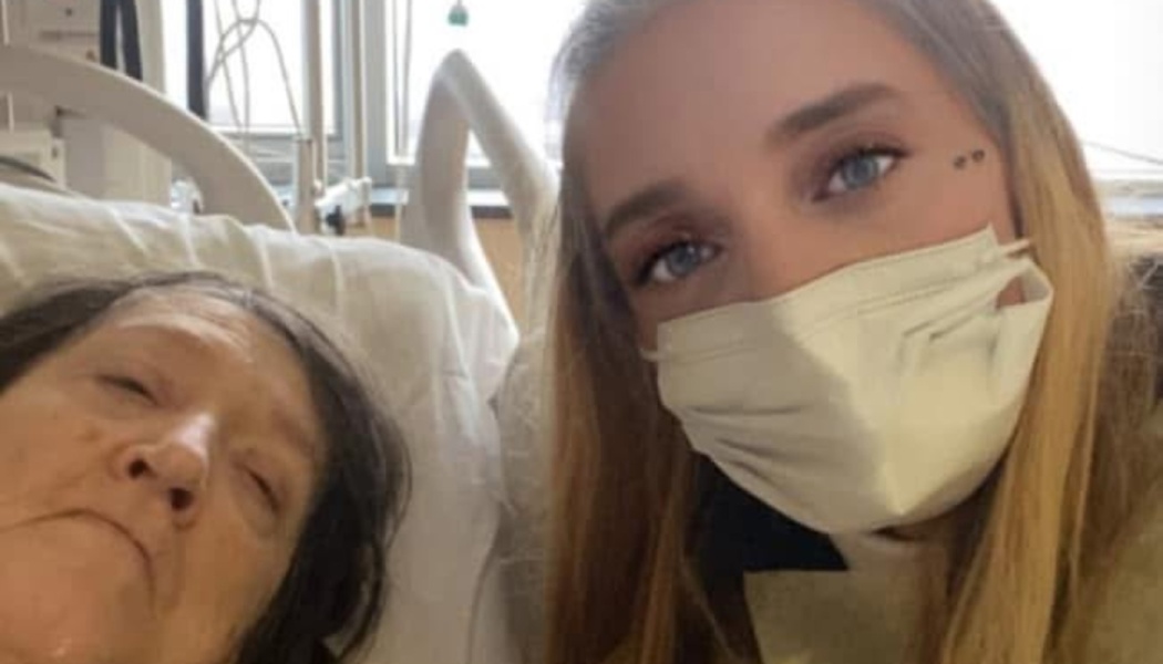 A young woman wearing a face mask takes a selfie next to an older woman resting in a hospital bed.
