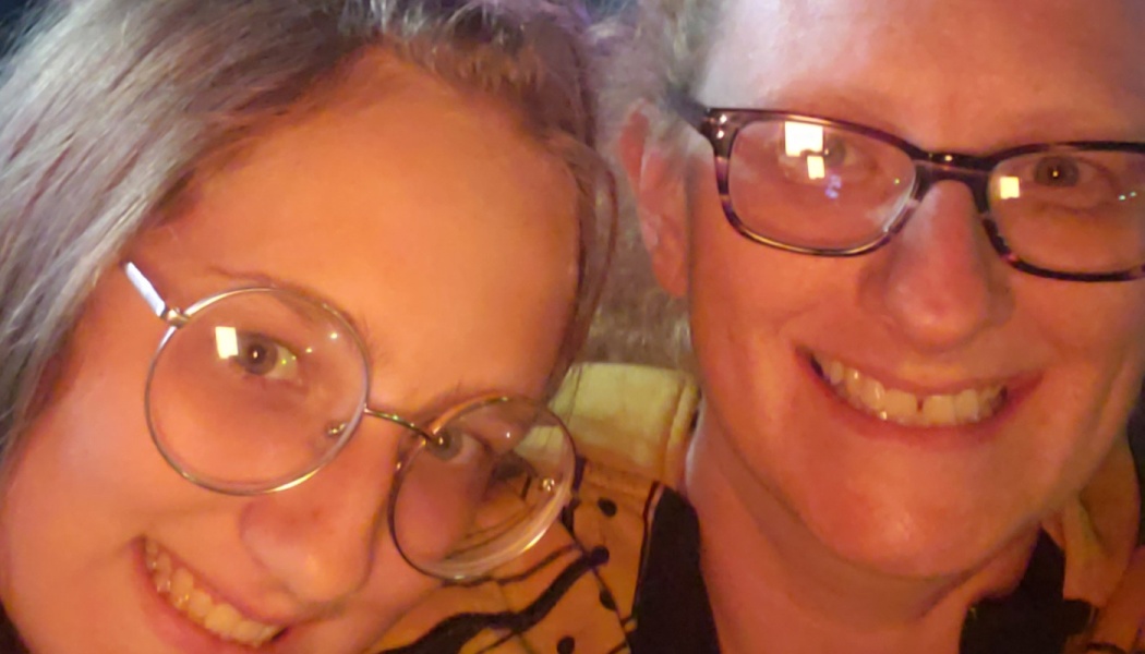 Close-up of two smiling people wearing glasses posing together in warm lighting.