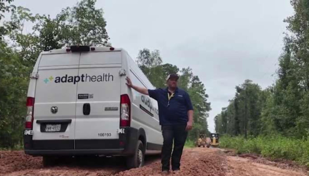 A man leans on a white AdaptHealth delivery van stuck on a muddy dirt road surrounded by trees.