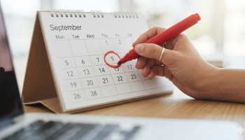 A hand uses a red marker to circle September 9th on a desktop calendar next to a laptop.