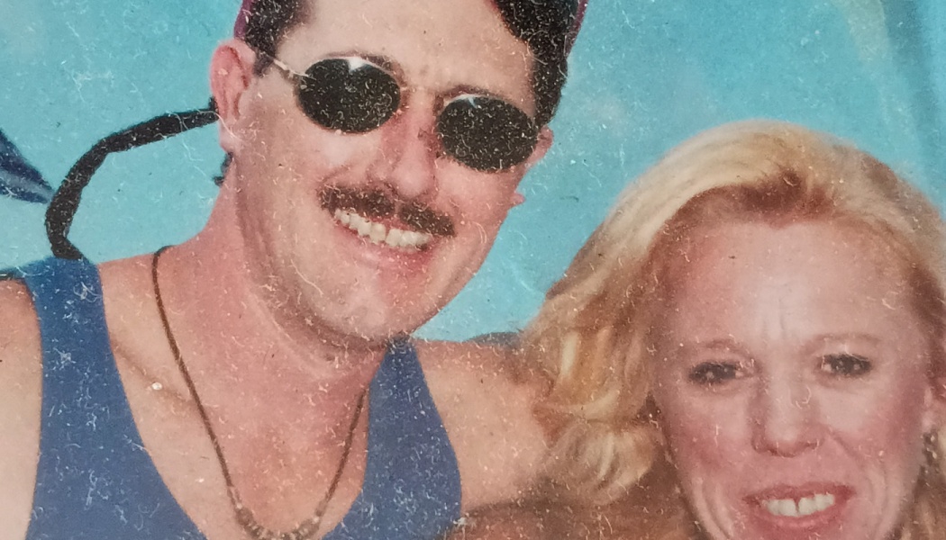 A smiling man with a mustache and dark sunglasses posing close to a smiling blonde woman.