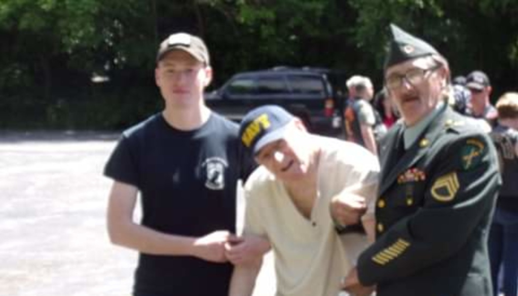 A young man and a uniformed Army veteran support an elderly Navy veteran standing outdoors.
