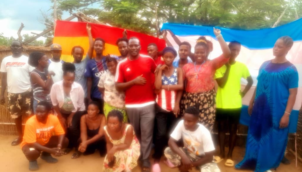A group of young adults gathered outdoors, holding up large rainbow and transgender pride flags behind them.