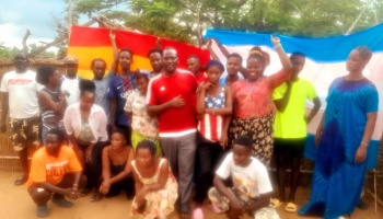 A group of young adults gathered outdoors, holding up large rainbow and transgender pride flags behind them.