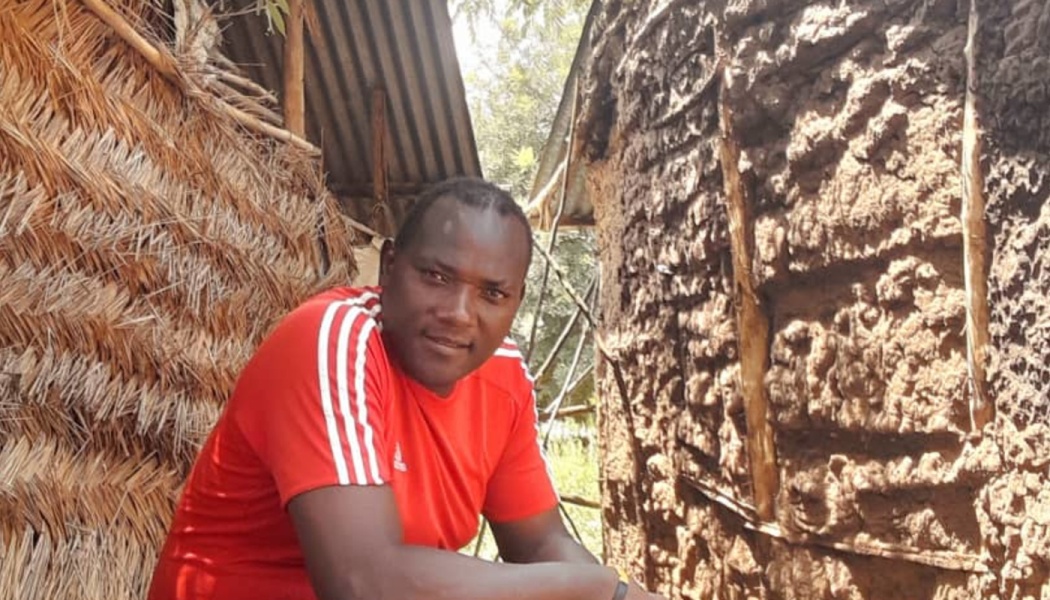 A man in a red t-shirt looks at the camera while leaning between a thatched hut and a mud wall.