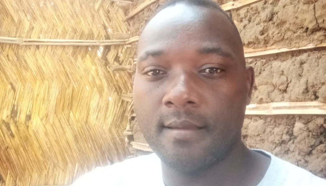 Close-up portrait of a man looking at the camera in front of a woven thatch and mud wall.