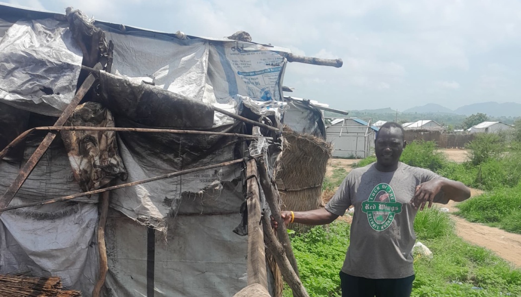 A man stands beside a fragile makeshift shelter made from worn tarps and wooden poles.