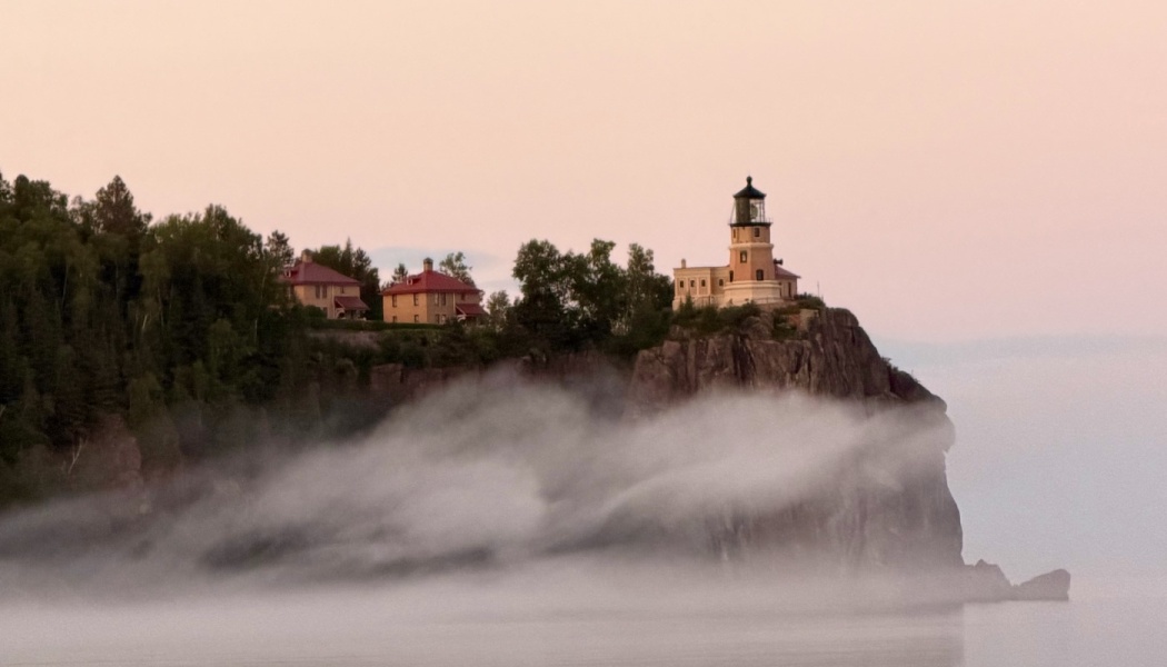 A historic lighthouse sits atop a high cliff surrounded by swirling fog under a soft pastel pink sky.