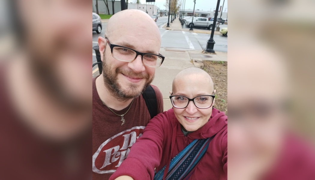 A smiling bald man with a beard and a smiling bald woman wearing glasses take a selfie outdoors.