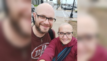 A smiling bald man with a beard and a smiling bald woman wearing glasses take a selfie outdoors.