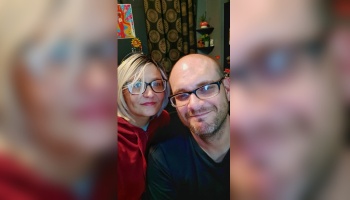 A smiling woman with short blonde hair and a bald man with a beard, both wearing black glasses, posing together.