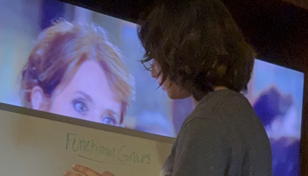 A dark-haired person stands near a whiteboard reading "Functional Grasps" in front of a large screen.