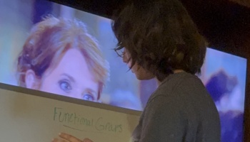 A dark-haired person stands near a whiteboard reading "Functional Grasps" in front of a large screen.