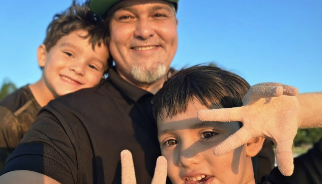 A smiling father poses outdoors with his two young sons, one showing off a sandy hand to the camera.