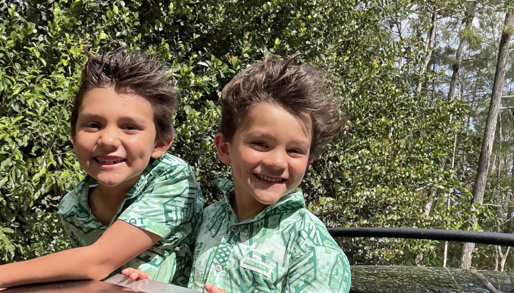Two young boys with windblown hair smiling in matching green shirts outdoors.