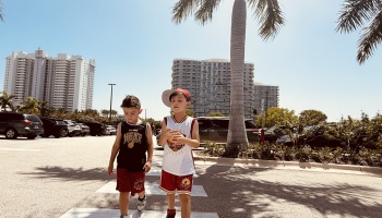 Two young boys wearing Miami Heat basketball jerseys walk outside on a sunny day near palm trees and tall buildings.