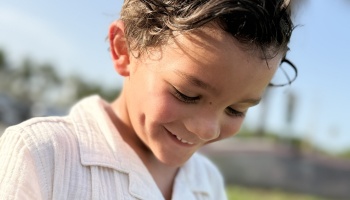Young boy with dark wavy hair and dimples looking down with a gentle smile outdoors in sunlight.